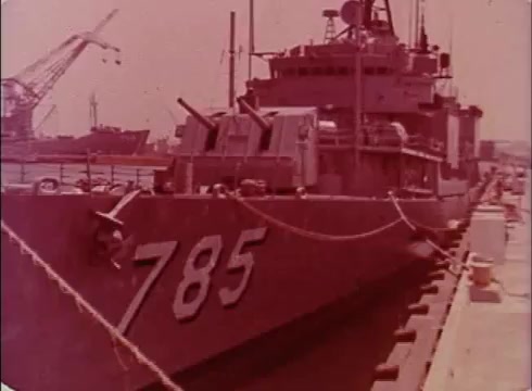 The still shows a military vessel identified by the number '785,' docked at a port. The ship appears to be a smaller craft, possibly involved in coastal defense or support roles, surrounded by ropes securing it to the dock. In the background, cranes and additional ships can be seen, suggesting a busy maritime environment. The color tone is reddish, indicative of the 1970s film style.