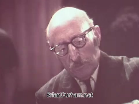 The 16mm still from 'Nabisco Escort Crackers - 1970s' features an older man with glasses and a mustache, dressed in a suit. He appears to be engaged in a thoughtful or serious task, possibly examining or discussing product details. The background is slightly blurred, suggesting a focus on the individual. The image captures a vintage aesthetic typical of the era.