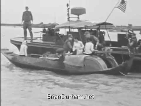 The still depicts a scene from the Vietnam era featuring military personnel on a boat alongside a smaller vessel. Soldiers are seen engaged with civilians on the water, suggesting a focus on community and support during the conflict. The presence of an American flag signifies a patriotic theme, likely aligning with the message of buying war bonds for supporting the troops.