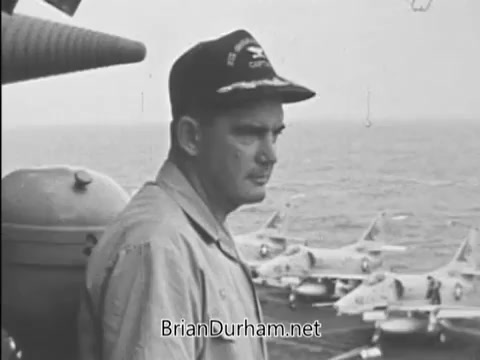 The still from 'Buy Bonds PSA - Vietnam Era - 1968' features a military officer on a naval ship, overlooking a flight deck with aircraft visible in the background. The officer, wearing a military cap and a buttoned shirt, gazes thoughtfully at the sea, conveying a sense of duty and contemplation during the Vietnam War era. The image is a part of a public service announcement aimed at encouraging support for war bonds.