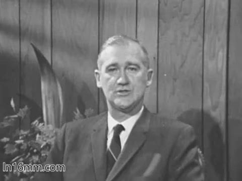 The still features a man in a suit and tie, seated against a wooden panel backdrop. He is likely part of a public service announcement (PSA) for the Veterans Administration from the 1960s. The setting appears formal and includes some greenery, suggesting a focus on service and support for veterans. The tone is serious, appropriate for the message being conveyed.