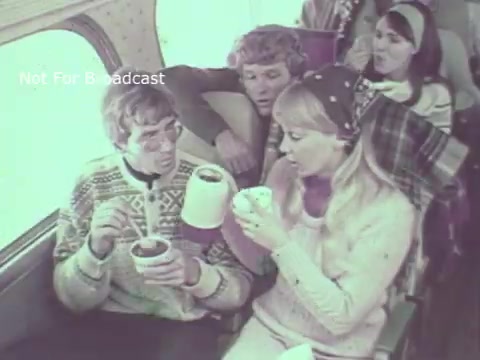 The still from the 1970 Hormel Chili TV commercial features a group of people seated on an airplane. Two of the individuals, a man and a woman, are enjoying bowls of chili, while another man and a woman are sitting nearby, seemingly engaged in a conversation. The overall mood appears casual and friendly, highlighting the enjoyment of Hormel Chili during travel. The image has a nostalgic quality, reflective of the era's style and advertising.