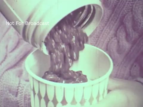 The still features a close-up of a hand pouring Hormel Chili from a container into a cup. The chili has a thick, hearty appearance and flows smoothly from the container. The background suggests a vintage setting, possibly from the 1970s, with a soft color tone typical of 16mm film. The overall feel evokes a casual, homestyle atmosphere, highlighting the product's appeal.