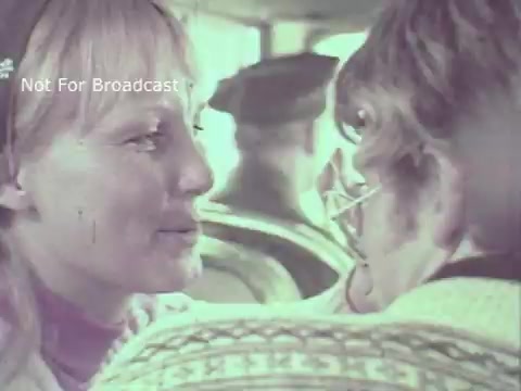 The still from the 1970 Hormel Chili TV commercial features a close-up of a woman with long blonde hair interacting with a man. They are positioned closely, likely engaging in a conversation. The background appears to show a bus or similar enclosed setting. The overall tone seems warm and lighthearted, reflecting the casual nature of the advertisement. The image captures a moment of connection and intimacy, fitting the playful theme of the commercial.