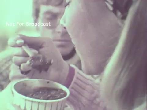 The still captures a scene from a 1970 Hormel Chili TV commercial, featuring a woman holding a cup of chili while taking a bite with a spoon. She appears engaged and enjoying the chili, with a male figure visible in the background, seemingly observing her. The overall aesthetic reflects the vintage style of the early '70s, possibly with a light color wash typical of that era’s film. The phrase “Not For Broadcast” suggests it's a production or promotional still rather than intended for public airing.