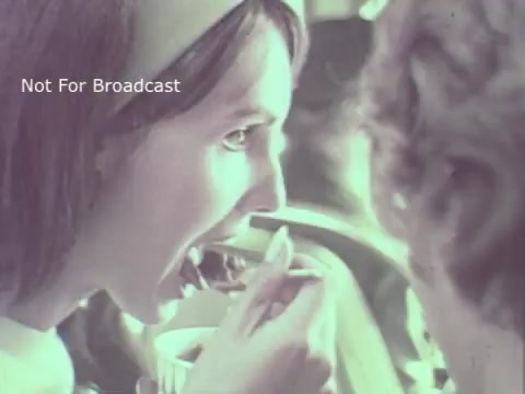 The still features a close-up of a woman taking a bite of food, likely from a spoon, in a setting illuminated by soft, warm lighting. Her expression suggests enjoyment or curiosity. The background shows blurred figures, adding to the cozy atmosphere of the scene. The overall aesthetic is characteristic of 1970s commercial style, with a nostalgic vibe. The image is marked 'Not For Broadcast.'