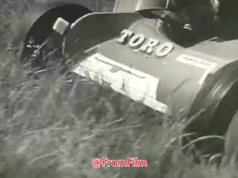 The still features a Toro Whirlwind lawnmower cutting through grass. The mower is predominantly in focus, showcasing its design and branding, with 'TORO' clearly visible. The background consists of tall grass being trimmed, capturing the essence of lawn care in the 1960s. The image is in black and white, typical of the era's commercial film style, conveying a nostalgic view of outdoor maintenance.