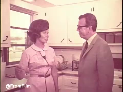 The still from the 'Cold Power Detergent TV Commercial' features a woman in a pink dress talking to a man in a suit, set in a brightly lit kitchen typical of the 1960s. The kitchen has wooden cabinets and a vintage aesthetic, reflecting the era's design. The woman's animated expression suggests she is sharing information about the detergent, while the man appears to be attentively listening. The overall scene captures the domestic vibe and marketing style of that time, focusing on household products.   Follow us on Twitter at @FromFilm.