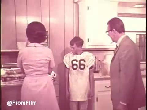 The still from the 'Cold Power Detergent TV Commercial' features a domestic kitchen scene typical of the 1960s. A woman in a light dress faces a kitchen appliance while a young boy in a football jersey stands nearby, looking slightly apprehensive. An older man, presumably a parent, watches from the side, engaged in the moment. This setup conveys themes of family life and everyday challenges with laundry, highlighting the product's effectiveness. The visual style reflects the era's color palette and fashion.   For more vintage insights, follow us on Twitter at @FromFilm.