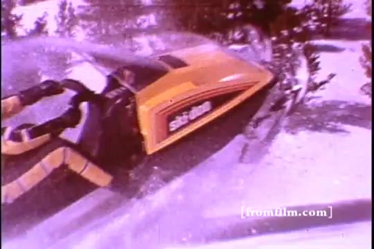 The still from the 1970s Ski-Doo snowmobile commercial features a close-up view of a bright yellow Ski-Doo snowmobile navigating through a snowy landscape. The snowmobile is shown in action, with snow being kicked up around it, emphasizing its speed and agility. The Ski-Doo logo is prominently displayed on the vehicle, showcasing its iconic design. The scene captures the essence of winter sports and the thrill of snowmobiling, appealing to adventure enthusiasts.