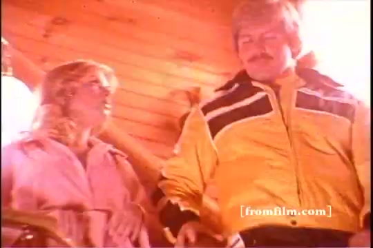 The still from the 'Ski-Doo' snowmobile commercial features two individuals inside a cozy log cabin setting. The man, wearing a bright yellow ski jacket with black accents, has a mustache and appears engaged in conversation with a woman dressed in a pink jacket. The warm, wooden interior suggests a winter sports theme, aligning with the commercial's focus on Ski-Doo snowmobiles and winter activities. The overall vibe is nostalgic, reflecting the spirit of the 1970s winter sports culture.