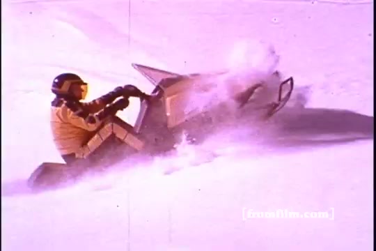 The still from the 1970s Ski-Doo snowmobile commercial features a snowmobiler in a vibrant yellow outfit riding a Ski-Doo snowmobile. The scene captures the excitement of winter sports, with the snowmobile kicking up snow as it maneuvers through a snowy landscape. The overall tone reflects the adventurous spirit of skiing and snowmobiling, characteristic of Ski-Doo's promotional style during that era.