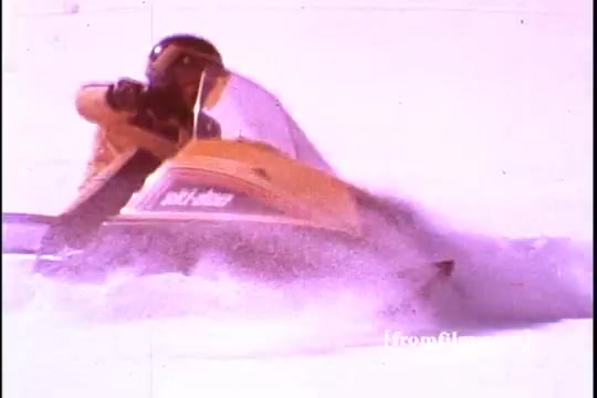 The still from the Ski-Doo snowmobile commercial features a vibrant yellow snowmobile cutting through fresh snow, leaving a spray of powder in its wake. The rider, dressed in winter gear, leans forward, indicating speed and excitement. The snowy landscape suggests a winter sports theme, capturing the essence of adventure associated with Ski-Doo snowmobiles, a well-known brand in Canadian winter recreation. The vintage feel highlights the commercial's 1970s era.
