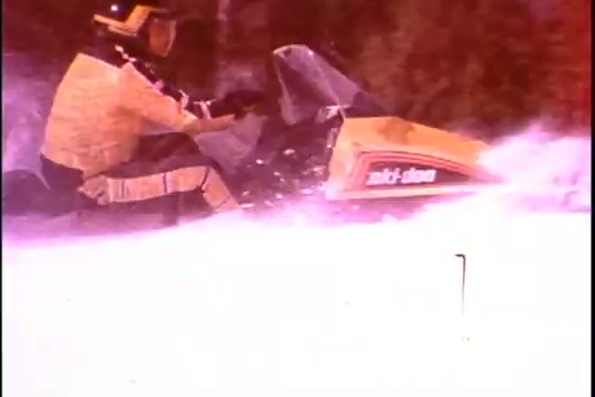 The still from the Ski-Doo snowmobile commercial features a rider in a bright yellow outfit maneuvering a snowmobile through a snowy landscape. The Ski-Doo model is prominently displayed, showcasing its design typical of the 1970s. The scene captures the thrill of winter sports, emphasizing the excitement and adventure associated with snowmobiling. The vibrant colors and action-oriented setting highlight the brand's appeal as a staple in winter recreational activities, particularly in Canada.
