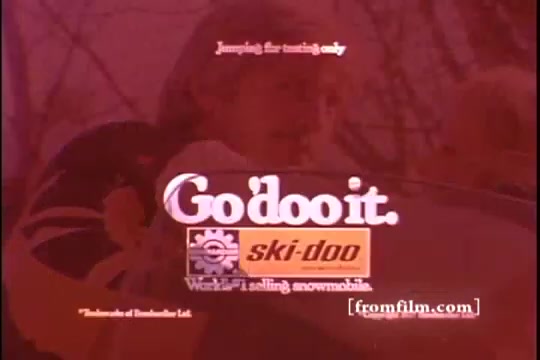The still features a vintage Ski-Doo commercial from the 1970s, prominently displaying the slogan 'Go'doo it' alongside the Ski-Doo logo. It suggests a focus on winter sports and snowmobiling, capturing the adventurous spirit associated with the brand. The background hints at a snowy outdoor setting, emphasizing the recreational aspect of Ski-Doo snowmobiles. The text includes a disclaimer about the jumping being for testing only, reinforcing safety while promoting the excitement of the product.