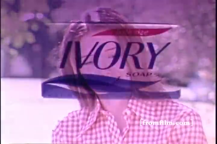 The still features a young woman smiling, wearing a checkered shirt. Overlaid prominently is the Ivory Soap logo, which includes the brand name and tagline. The background is soft-focused, creating a nostalgic and warm atmosphere typical of 1970s advertisements. The color palette is muted, in line with the era's aesthetic.