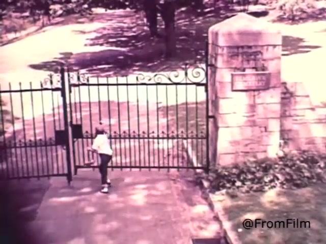 The still from 'Sealmark Gravestone Markers - 1968' features a child approaching a wrought iron gate. The background suggests a serene, possibly cemetery-like setting, with well-maintained grounds. The gate is partially open, inviting exploration. Accompanying voice-over highlights the significance of selecting a reliable figure in the community for memorial services, emphasizing the trustworthiness of Sealmark as a brand. The overall tone reflects a respectful acknowledgment of life's transitions.