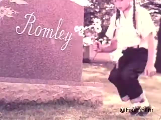 The 16mm still from 'Sealmark Gravestone Markers - 1968' features a young child with long braids, dressed in a white shirt and black pants, kneeling beside a gravestone marked 'Romley.' The scene is set in a serene outdoor environment, suggesting a moment of remembrance. The child's gesture or interaction with the gravestone adds a poignant touch to the advertisement's message about the importance of remembering loved ones and choosing a respected name in monuments.