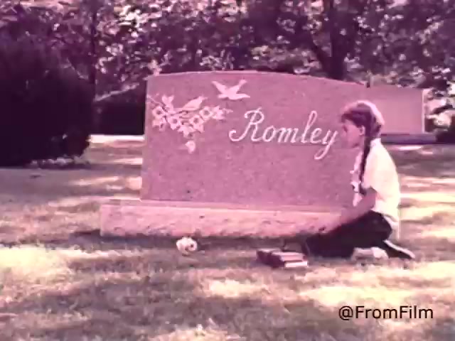 The still depicts a young girl kneeling in front of a gravestone marked 'Romley,' surrounded by a serene cemetery setting. The gravestone features decorative engravings of birds and flowers. The girl is holding some items, possibly flowers or mementos, conveying a sense of remembrance and reflection. The background is lush with greenery, adding to the tranquil atmosphere. The voice-over emphasizes the reliability of Sealmark as a trusted provider of memorials during difficult times.
