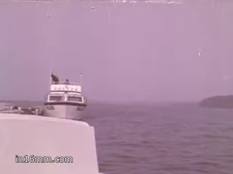 The 16mm still depicts a scene from a 1980s boating safety PSA produced by NOAA. It features a boat navigating on calm waters, with a visible shoreline in the background. The color palette is soft, typical of that era's film quality. This PSA likely emphasizes the importance of boating safety and the use of NOAA Weather Radio for marine communication and alerts.