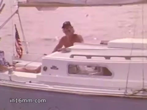 The still from the 16mm PSA features a man on a sailboat, with an American flag visible in the background. He appears relaxed, possibly enjoying a sunny day on the water. The focus of the PSA is on promoting boating safety and the importance of NOAA Weather Radio for mariners, likely to emphasize awareness of weather conditions while boating. The scene conveys a sense of leisure, set against the backdrop of a calm waterway.