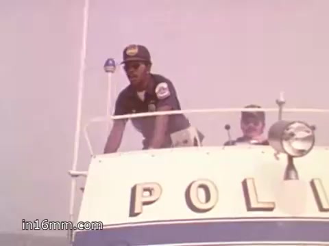 The still shows two figures on a police boat, presumably part of a boating safety campaign. One individual, wearing a police uniform and sunglasses, is at the helm, looking out over the water. Another person is seated behind him. The boat's side prominently displays the word 'POLICE.' The scene is likely emphasizing safety and awareness in boating activities. The image reflects a time when public service announcements were aimed at educating the public on marine safety.