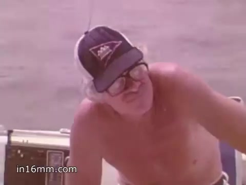 The still shows an older man on a boat, wearing glasses and a cap with a logo. He appears to be focused or contemplative, possibly engaging in a discussion about boating safety. The background suggests a water setting, aligning with the theme of the PSA promoting NOAA Weather Radio for safe boating practices. The image captures a moment that conveys the importance of awareness and preparedness while on the water.