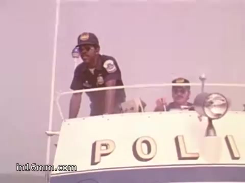 The still from the 16mm PSA shows two police officers aboard a boat, likely emphasizing boating safety. One officer is standing at the helm, gazing intently ahead, while the other is seated beside him, possibly engaged in conversation. The boat is marked 'POLICE,' indicating it’s a patrol or safety enforcement vessel. The setting appears to be on water, with a calm atmosphere. This image reflects the PSA's focus on promoting safe boating practices, likely in connection with NOAA Weather Radio information.