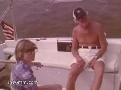 The still from the 1980s boating safety PSA features two men aboard a sailboat. One man, wearing glasses and a plaid shirt, appears to be engaged in conversation while looking towards the camera. The other, a shirtless man with a cap, sits relaxed with his legs crossed. The boat's interior is simple, with an American flag visible in the background, and the water can be seen behind them. The image reflects a casual moment while promoting boating safety awareness.