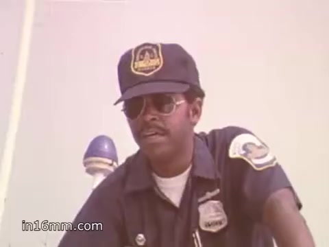 The still features a man wearing a police-style uniform and sunglasses, with a badge visible on his shirt. He appears to be engaged in a conversation or providing information, likely emphasizing the importance of boating safety. The background is light-colored, adding to a bright, outdoor vibe typical of the 1980s. This still is part of a 30-second public service announcement promoting NOAA Weather Radio for boating safety.