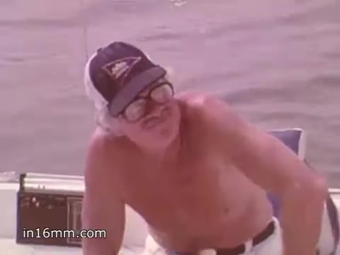 The still from the 16mm PSA titled 'Boating Safety - NOAA Weather Radio' captures a middle-aged man on a boat, wearing a baseball cap and large glasses. He appears shirtless, suggesting a casual boating environment. The background features water, indicating the boating context. The PSA likely promotes safe boating practices and the importance of NOAA Weather Radio for weather updates. The shot is in color, typical of the 1980s, and conveys a relaxed yet informative tone.