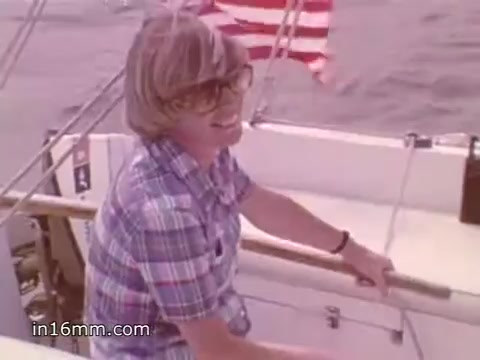 The still from the 16mm PSA titled '1980's Boating Safety - NOAA Weather Radio' depicts a person on a boat, wearing a plaid shirt and large glasses, smiling while holding onto a boat's railing. In the background, there is an American flag fluttering against a backdrop of water. The overall scene evokes a sense of leisure and safety on the water, aligning with the PSA's focus on boating safety and the importance of NOAA Weather Radio for sailors.