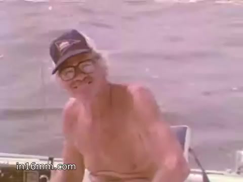 The still shows an older man with glasses and a cap, smiling and looking directly at the camera. He is shirtless and situated on a small boat, with the water visible in the background. The scene conveys a relaxed, boating atmosphere, likely promoting safety on the water, emphasizing the importance of NOAA Weather Radio in a playful or approachable manner.