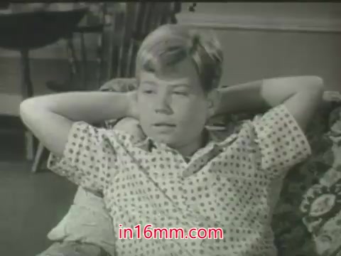 The 16mm still from the ABC promo of 'My Three Sons' features a young boy lounging on a couch, appearing relaxed with his hands behind his head. He is dressed in a patterned short-sleeve shirt typical of the 1960s. The black-and-white format emphasizes the classic television aesthetic of the era. The background likely includes elements of a cozy home setting, typical of the show's family-oriented themes. The presence of William Frawley indicates the time frame aligns with the series' early seasons on ABC, before its transition to CBS.