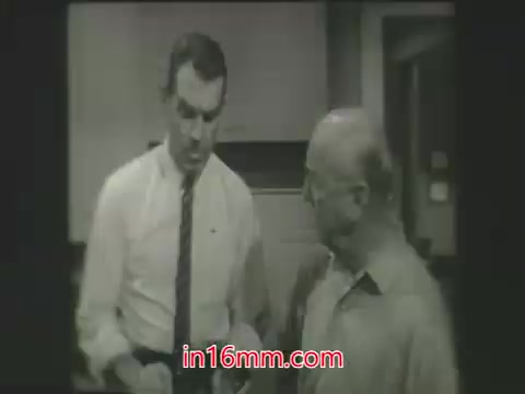 The still from 'My Three Sons' features two characters engaged in a conversation. One man, dressed in a white shirt and tie, appears to be younger and is discussing something with an older man, who is bald and wearing a button-up shirt. The scene captures a moment of interaction between the characters, set in a domestic or office environment typical of the series. The black and white format emphasizes the classic television aesthetic of the 1960s.
