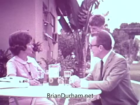 The still features a scene set outdoors, likely in a Florida backyard. A woman in a light-colored outfit engages in conversation with a man in a suit, while a younger boy stands nearby. They are gathered around a table with drinks and a box of Cold Power Detergent visibly placed on it. The lush greenery and casual setting suggest a relaxed, familial atmosphere.