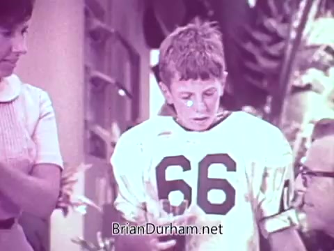 The still features a young boy wearing a football jersey with the number 66, appearing slightly dirty, which may imply he just finished playing. He is surrounded by adults, including a woman and a man wearing glasses. They look on as he holds a drink, possibly indicating a post-game scene. The setting appears casual and domestic, reflecting a light-hearted family moment. The commercial promotes Cold Power Detergent, likely emphasizing its cleaning effectiveness after sports activities.