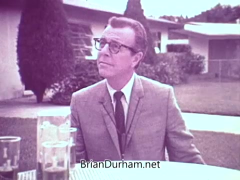 The still features a well-dressed man in a suit, seated outdoors at a table, possibly promoting Cold Power Detergent. The setting includes green landscaping typical of Florida, indicating a warm, relaxed atmosphere. The man's expression suggests he is engaged in conversation or making a point about the detergent’s effectiveness. In the foreground, there are glasses, likely of a beverage, adding to the casual vibe of the scene.