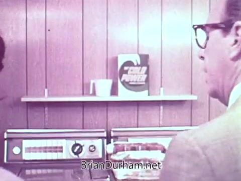 The still features two men in a kitchen setting, facing a vintage washing machine. On the shelf behind them is a box of Cold Power detergent prominently displayed. The decor has a retro look, with wooden paneling and appliances typical of the era. The scene suggests a domestic or promotional context, highlighting the product's appeal.