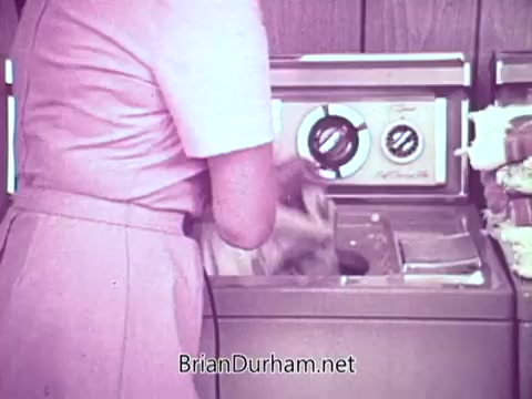 The still features a woman in a pink dress loading laundry into a washing machine. The machine is vintage, with dials for settings visible. The background suggests a home environment, likely a kitchen or laundry room, emphasizing a domestic setting. The image captures a typical moment of laundry day, promoting the practical use of Cold Power Detergent.