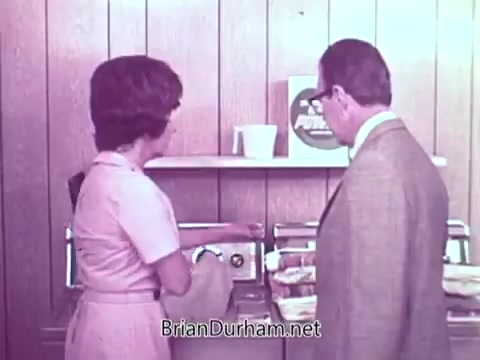 The still features a woman in a pink dress and a man in formal attire standing near a countertop. The woman is interacting with a washing machine or laundry appliance, while the man observes. The setting has a retro aesthetic, characterized by wooden paneling and a visible detergent box labeled 'Cold Power.' The overall tone of the image suggests a domestic scene focused on laundry.