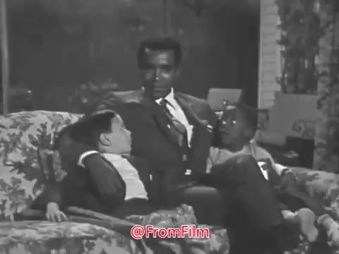 The still from 'Greg Morris PSA for Easter Seals' features Greg Morris seated on a couch, interacting with two young boys. The scene exudes a warm, family-oriented atmosphere, with Morris engaging in a conversation that highlights his empathetic demeanor. The setting appears cozy, likely intended to convey a message of support and connection, aligning with the themes of the Easter Seals organization.
