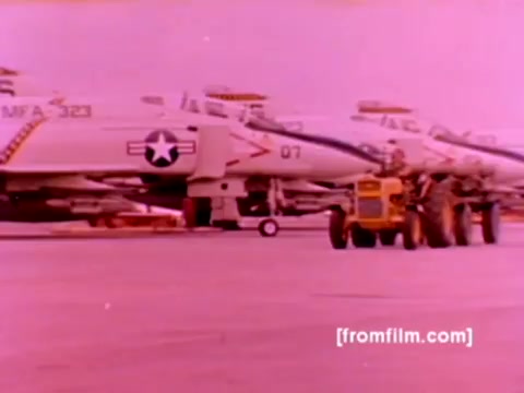 The still from the 'USMC Aviation Ordnance enlistment film' features a lineup of F-4 Phantoms, AH-1 SuperCobras, and an FA-18, with a Harrier also included in the context of ordnance operations. In this 16mm negative, aircraft are prominently displayed on the tarmac, being prepared for ordnance loading, showcasing the process of mounting missiles and bombs. The color has a pronounced pink hue typical of vintage film stock, indicating a need for color correction for clarity and realism. The aircraft are positioned with ground support equipment visible, emphasizing the operational environment of Marine Corps aviation.