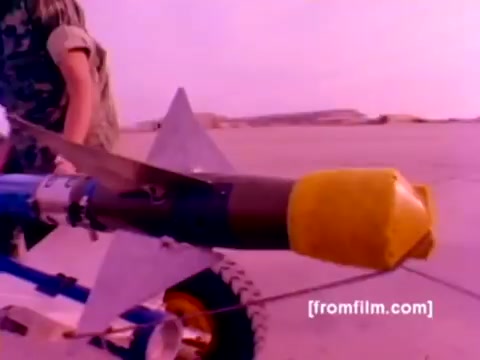 The still from the 'USMC Aviation Ordnance enlistment film' showcases a close-up view of an ordnance device being prepared for mounting. The ordnance features a distinct conical nose, painted yellow, indicating it is likely a missile or bomb. In the background, a military personnel in camouflage attire is involved in the process, contributing to the hands-on depiction of ordnance handling within the Marine Corps.  The overall color palette appears to be enhanced with a pinkish hue, likely due to the 16mm film's vintage origins and the color correction needed for clarity. The still encapsulates the focus on aviation ordnance, illustrating the technical and operational elements of the Marine Corps’ aviation capabilities.