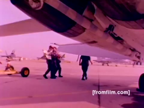 The still from the 'USMC Aviation Ordnance' enlistment film captures a moment on a military airfield, showcasing F-4 Phantoms, AH-1 SuperCobras, an FA-18, and a Harrier in the background. In the foreground, personnel are seen meticulously mounting ordnance, including missiles and bombs, under the wing of an aircraft. The image, derived from a 16mm negative, has a muted color palette, typical of vintage films, likely needing enhanced color correction for clarity. The scene emphasizes the technical and operational aspects of Marine Corps aviation ordnance responsibilities.