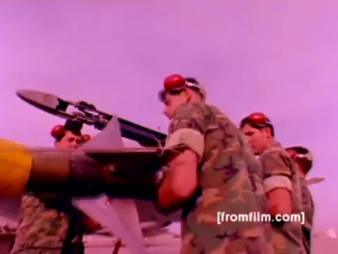 The still from the USMC Aviation Ordnance enlistment film features a group of servicemen in camouflage uniforms, wearing red headsets as they work together to mount ordnance, specifically a missile, onto military aircraft. In the background, F-4 Phantoms, AH-1 SuperCobras, an FA-18, and a Harrier are visible, showcasing the impressive array of Marine Corps aviation assets. The scene conveys a sense of teamwork and precision involved in ordnance handling, emphasizing the technical aspects of military service in aviation. The color correction would enhance the vibrancy of the uniforms, aircraft, and ordnance, giving a more accurate representation of the environment.