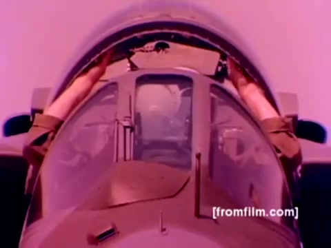 The still from the 'USMC Aviation Ordnance enlistment film' captures a close-up view of the cockpit of an aircraft, likely an F-4 Phantom or similar model. Two hands are visible, working on the cockpit controls or instruments, emphasizing the technical aspects of aviation ordnance tasks. The background hints at a purple hue, indicative of the film’s age and original color grading from the 16mm negative.  In this film, the focus is on the ordnance option for enlistment, showcasing the Marine Corps' sophisticated aircraft like F-4 Phantoms, AH-1 SuperCobras, FA-18s, and Harriers, while highlighting the mounting of missiles and bombs during pre-flight preparations. The image evokes a sense