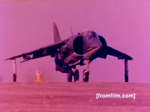 The still from the 'USMC Aviation Ordnance enlistment film' captures a Harrier jet preparing for takeoff on a runway. The Harrier is prominently featured in the foreground with its wings in a slightly angled position, showcasing its unique vertical takeoff capability. The background appears to be a blurred runway or ramp area. The color correction reveals an overall pinkish hue, possibly due to the 16mm film's age and processing. This image aligns with the film's focus on ordnance and aircraft, highlighting the enlistment option within the Marine Corps aviation sector.