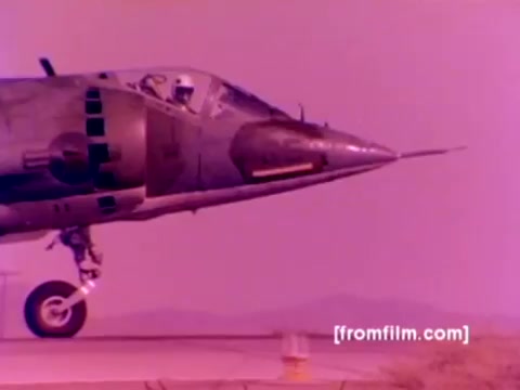 The still captures a close-up view of a Harrier jet, showcasing its distinctive nose and cockpit design. The aircraft is positioned as if preparing for takeoff, with a blurred background that suggests an airfield or open terrain. The film’s color has a faint pinkish hue typical of older 16mm footage but can be enhanced for clarity. Key elements include the detailed texture of the jet's surface and the pilot visible in the cockpit. This image is part of a Marine Corps film emphasizing the 'Ordnance Option' in enlistment, featuring various military aircraft and ordnance tasks.