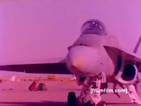 The still from the 'USMC Aviation Ordnance enlistment film' features an FA-18 aircraft prominently in the foreground. The image, originally from a 16mm negative, is characterized by a color cast that gives it a pinkish hue. The aircraft’s sleek design and notable features, such as the cockpit and landing gear, are clearly visible. In the background, other military equipment and possibly additional aircraft can be seen, suggesting an airfield setting. Overall, the still captures the essence of Marine Corps aviation and the ordnance operations involved.